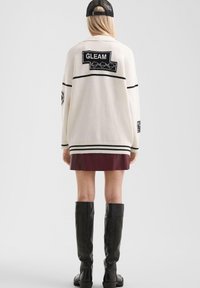 Kobieta w kremowym swetrze z naszywką "GLEAM", czarnym wykończeniem, bordowej spódnicy, czarnych wysokich kozakach i czapce z siateczki, stojąca tyłem.