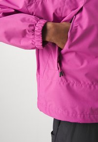 Main glissée dans la poche zippée d'une veste rose vif avec manches à poignets élastiques, portée sur un pantalon noir.