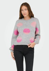 Femme aux longs cheveux bruns portant un pull gris à grands pois roses et oranges et un pantalon noir, se tenant devant un fond blanc.