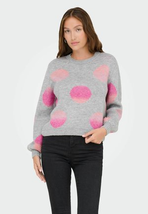 Frau mit langen braunen Haaren, die einen grauen Pullover mit großen rosa und orangefarbenen Punkten und schwarze Hosen trägt, steht vor einem weißen Hintergrund.