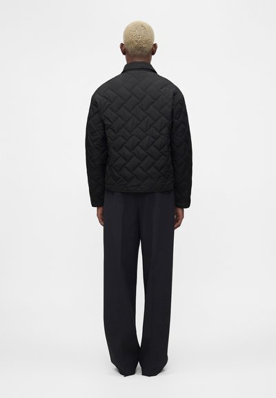 Άτομο με κοντά ξανθά μαλλιά, φαίνεται από πίσω, φοράει μαύρο καπιτονέ μπουφάν και φαρδιά μαύρα παντελόνια, στέκεται μπροστά σε απλό λευκό φόντο.