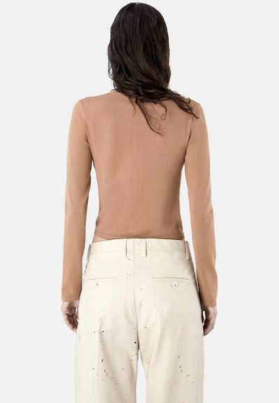 Femme aux longs cheveux foncés portant un haut moulant à manches longues de couleur camel et un pantalon ample beige clair avec des taches foncées, vue de dos.