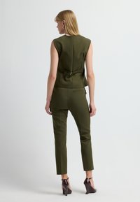 Zielono-oliwkowy, dopasowany garnitur z bezrękawnym topem z subtelnym wiązaniem z tyłu oraz wąskimi spodniami, zestawiony z połyskującymi ciemnymi szpilkami.