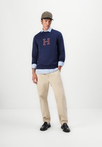 Felpa della marina con logo "H", indossata sopra una camicia a righe blu chiaro, pantaloni beige e scarpe nere. Cappello a scacchi con visiera curva.