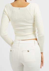 Krämfärgad ribbad långärmad topp med kvadratisk halsringning, kombinerad med vita jeans med slitna bakfickor och metalliska knappdetaljer.