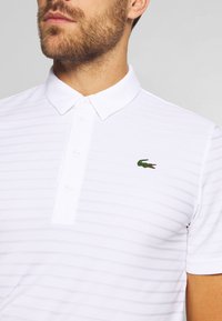 Valkoinen polo-paita, jossa on huomaamattomat pystyrivit, kolmella napilla varustettu kaulus ja vasemmassa rinnassa vihreä krokotiililogo. Pehmeä, textuuroitu kangas.