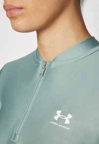 Šviesiai žalia zip-up sportinė palaidinė su apvaliu apykakle ir baltu Under Armour logotipu kairėje pusėje. Lygus, tamprus audinys.