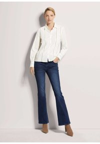 Witte button-up shirt met verticale kantdetails, gecombineerd met donkerblauwe flared jeans en tan puntige hakken. Gladde textuur, slim fit.