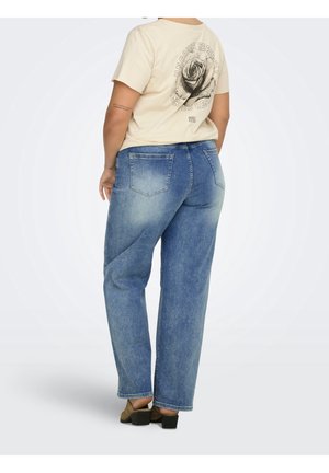 Person iført en lysebrun t-shirt med rosenmotiv og tekst, kombineret med blåslidte jeans og brune sko med blokhæl, stående med ryggen mod.