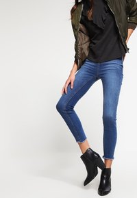 Ciemnozielona kurtka typu bomber na czarnej, falbaniastej bluzce, zestawiona z niebieskimi obcisłymi dżinsami i czarnymi botkami za kostkę. Gładki denim, detal z postrzępionym wykończeniem.