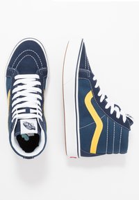 Vysoké tenisky s tmavě modrou suedinou, bílými šnami, žlutým bočním pruhem a gumovou podrážkou. Výrazné detaily šití na svršku.