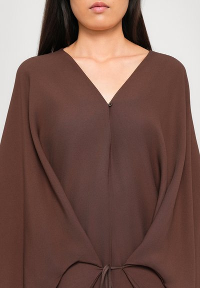 Woman wearing a loose brown V-neck top with a single button and a tied drawstring detail at the front center.