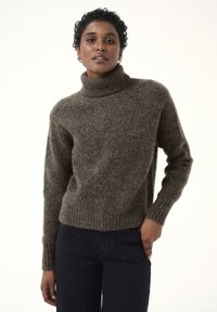 Brauner Strick-Turtleneck-Pullover mit gerippten Bündchen und Saum, der ein strukturiertes Finish aufweist. Kombiniert mit dunklen Jeans.