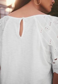 Un haut blanc avec un dos en trou de serrure et une fermeture à bouton, agrémenté de dentelle brodée sur les manches, fabriqué dans un tissu léger et texturé.