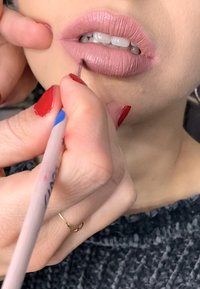 Lepapplikasjon i en myk rosa tone, med en leppestiftblyant som brukes; røde negler og en gullring er synlige. Teksturert mørk bakgrunn.