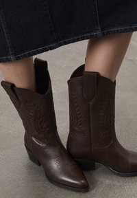 Bottes western en cuir marron avec des coutures détaillées, portées avec une jupe en denim foncé sur un sol gris texturé.