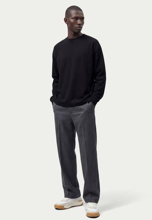 Man staat met handen in zakken, draagt een zwart shirt met lange mouwen, grijze broek en witte sneakers met gumzolen.