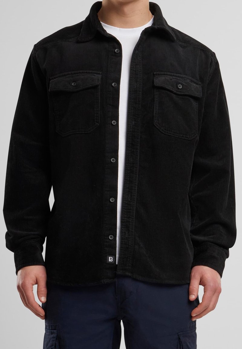 Man i svart kordflorsskjorta med knappar över en vit t-shirt, iförd mörkblå cargobyxor, står mot en enkel bakgrund.