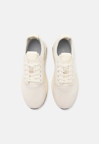 Light beige athletic shoes with a textured knit upper, flat laces, and a cushioned sole. Features a subtle logo near the tongue.