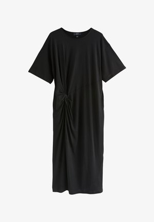 Femme aux cheveux bruns attachés en chignon, portant une robe midi noire à manches courtes avec un nœud devant, debout contre un mur clair texturé, chaussée de sandales noires.