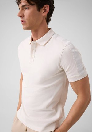 Junger Mann trägt ein schlichtes weißes Poloshirt mit kurzen Ärmeln, Kragen und Knopfleiste, steht mit den Händen in den Taschen.
