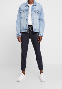 Person som bär en ljusblå denimjacka, en vit tryckt T-shirt, svarta joggerbyxor med dragkedjor och vita sneakers som står framför en enkel bakgrund.