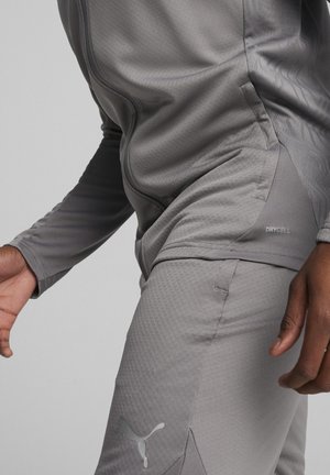 Das graue Sportoberteil und die Hose bestehen aus einem strukturierten Stoff mit einem figurbetonten Design. Das Oberteil hat einen Reißverschluss und Mesh-Akzente, und die Hose hat ein Logodetail.