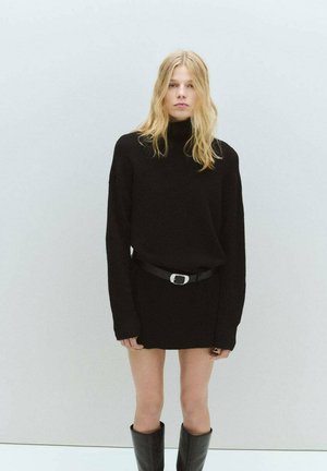 Vestido suéter negro de punto con cuello alto, mangas largas y cinturón en la cintura. Combinado con botas negras hasta la rodilla. Textura de tela suave.