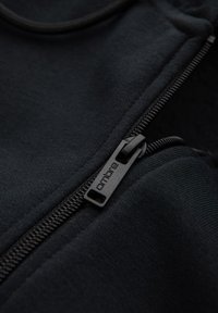 Gros plan sur un tissu noir avec une fermeture éclair noire partiellement dézippée, étiquetée « ombre » sur la tirette.