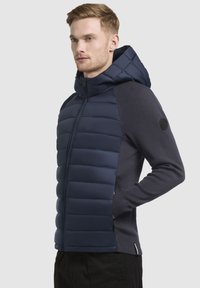Doudoune navy avec fermeture éclair à l'avant et capuche ; présente des sections rembourrées et des manches en tricot ; poches latérales ; texture de tissu lisse.
