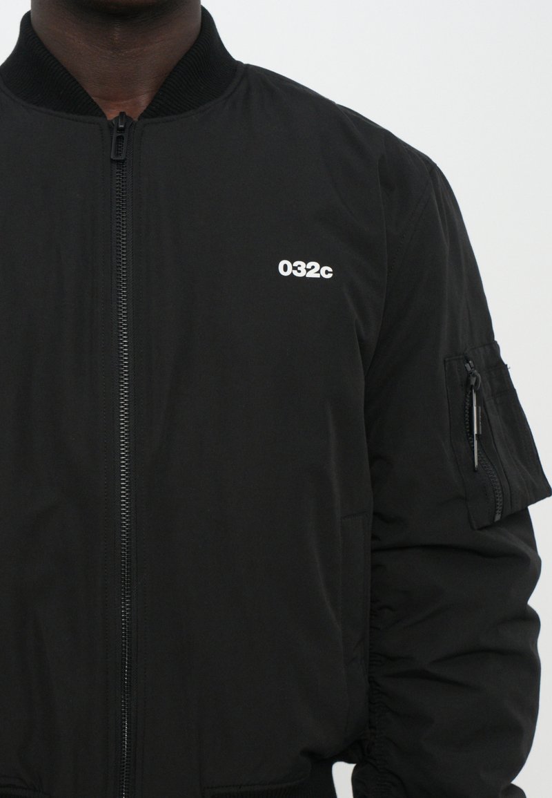 Chaqueta bomber negra con cremallera frontal, cuello y puños acanalados. Logo "032c" en blanco en el lado izquierdo del pecho y un bolsillo con cremallera en la manga.