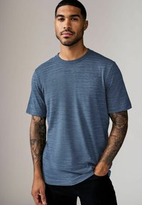Tricou albastru cu mânecă scurtă, cu un model texturat, decolteu rotund și croială lejeră, purtat cu pantaloni negri. Prezintă tatuaje vizibile pe brațe.