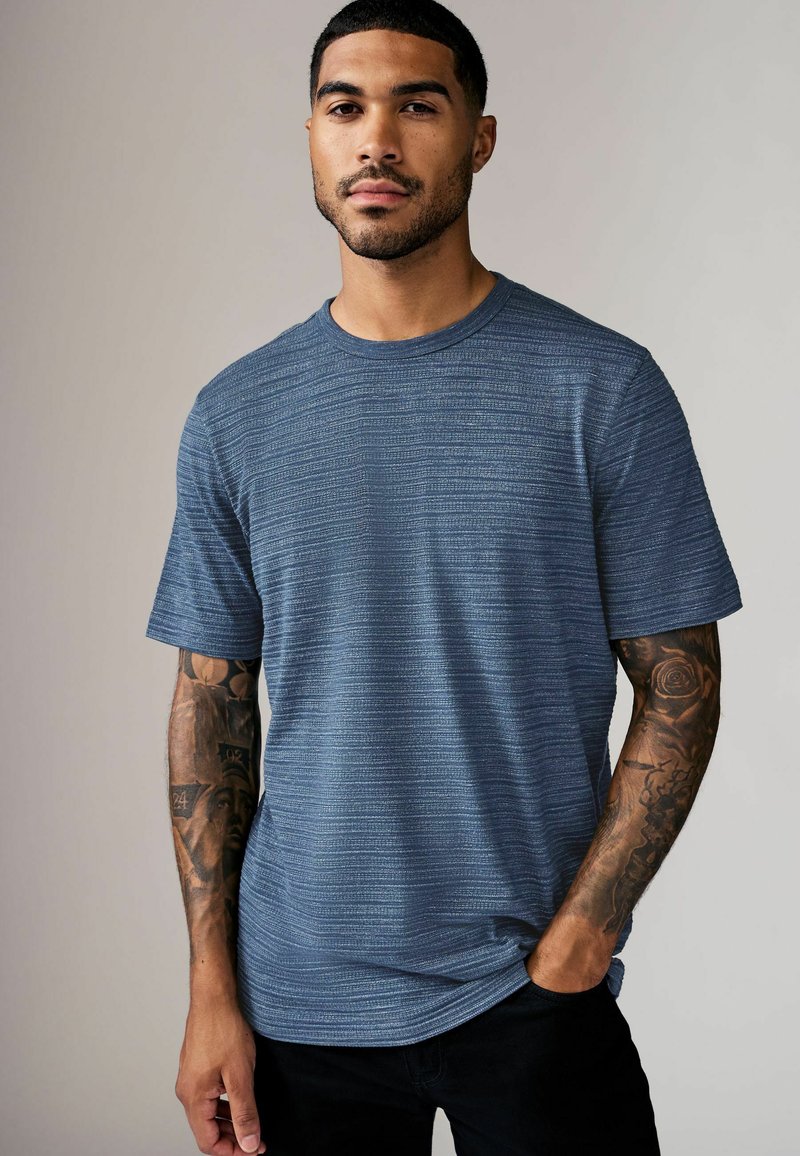 Tricou albastru cu mânecă scurtă, cu un model texturat, decolteu rotund și croială lejeră, purtat cu pantaloni negri. Prezintă tatuaje vizibile pe brațe.