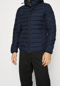 Mann trägt eine dunkelblaue gesteppten Daunenjacke mit Kapuze und Frontreißverschluss, kombiniert mit schwarzen Hosen, vor einem einfarbigen Hintergrund.