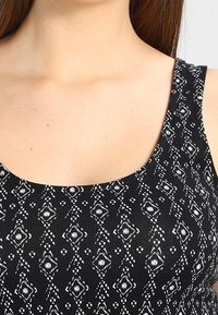 Černý top bez rukávů se bílým geometrickým vzorem obsahujícím diamanty a tečky, kulatým výstřihem a hladkou texturou látky.