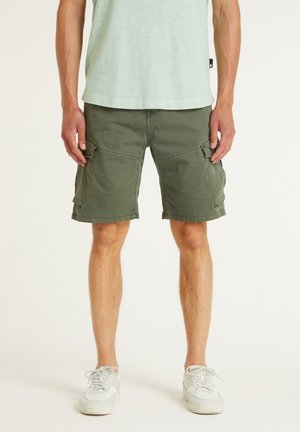 Man wearing olive green cargo shorts, white sneakers, and a light green shirt, standing against a plain background.