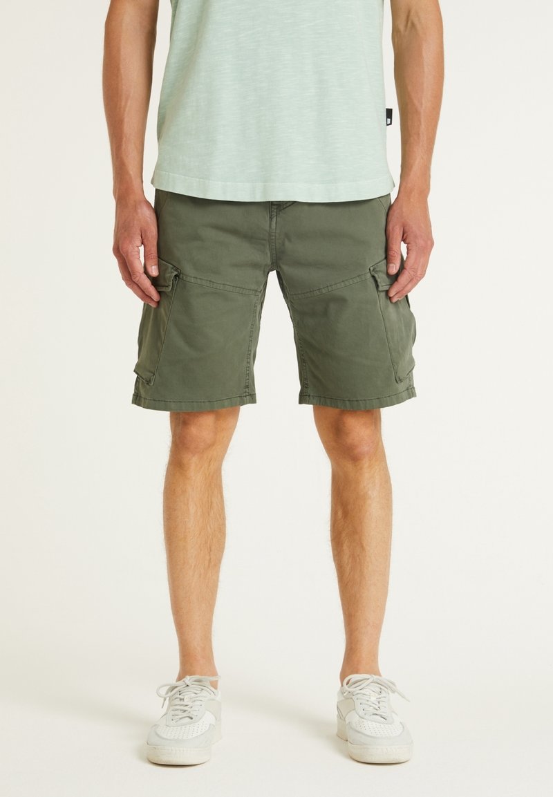 Homme portant un short cargo vert olive, des baskets blanches et une chemise vert pâle, debout devant un fond uni.