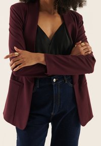 Burgunderfärgad kavaj med skräddarsydd passform, med framfickor och långa ärmar, bärs över en svart satin topp och högmidjade denimjeans.