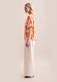 Kleurrijke bloemetjesblouse in oranje en roze, met pofmouwen; gecombineerd met witte, op maat gemaakte broek en zwarte, hoge sandalen.