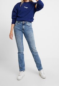 Sudadera azul con el logo de "Wrangler" y acentos de rayas multicolor en las mangas, combinada con jeans de pierna recta de color azul claro y zapatillas blancas.
