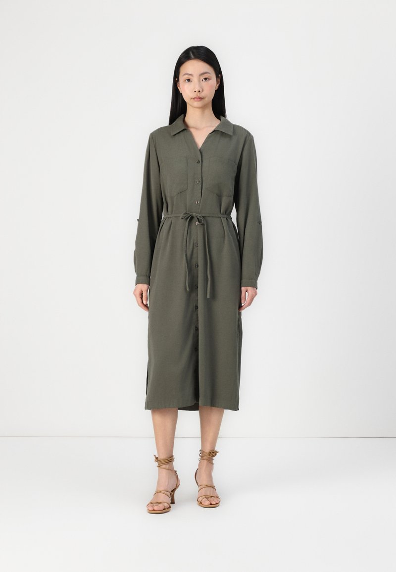 Vestido verde-oliva de mangas longas com fecho de botões, gola e cintura com laço. Apresenta dois bolsos no peito e rachas laterais. Usado com sandálias de tiras.