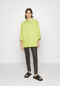 Camisa oversized verde-claro com colarinho pontudo e dois bolsos no peito, combinada com calças escuras de corte justo e sandálias pretas deslizantes.