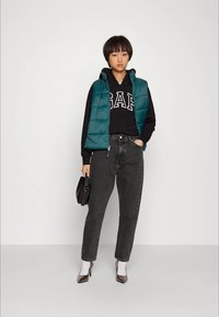 Jovem vestindo um colete acolchoado teal, hoodie preto da GAP, jeans escuros, meias brancas com calcanhares escuros, segurando uma pequena bolsa preta, de pé contra uma parede branca.