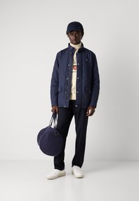 Námořnická prošívaná bunda, krémová mikina, tmavé kalhoty, bílé tenisky a kulatá námořnická taška. Texturovaná látka a kontrastní barvy.