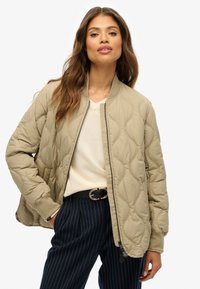 Beige gewatteerde bomberjack met een ritssluiting, geribde manchetten en een gladde textuur. Draag het over een lichte trui en een gestreepte broek.
