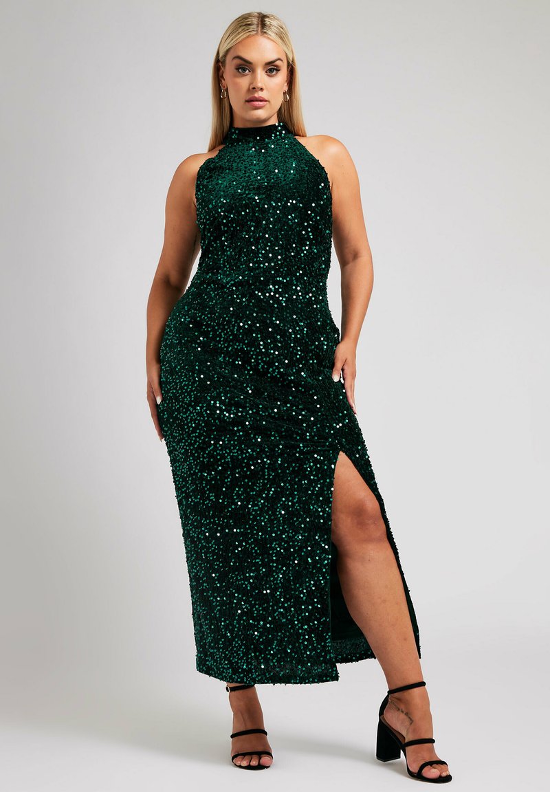 Grön paljettklänning med hög ringning, åtsittande form och hög slits vid låret. Har en mjuk textur och glänser starkt. Svarta sandaler med remmar.