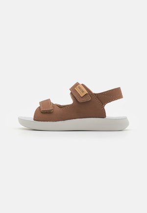Sandal de cuero marrón con dos correas de velcro, suela de goma blanca y etiqueta con el logo de Geox. Presenta un diseño abierto y detalles de costura.