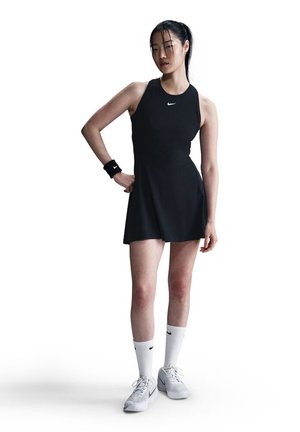 Fekete sportos ruha, testhez simuló felső résszel és bővülő szoknyával, fehér Nike zoknival és cipővel párosítva, valamint fekete karkötővel. Figyelemre méltó Nike logóval.