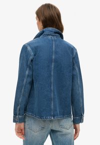 Chaqueta de denim azul con cuello estructurado, costuras visibles y detalles de puños con botón. La tela tiene un acabado texturizado.