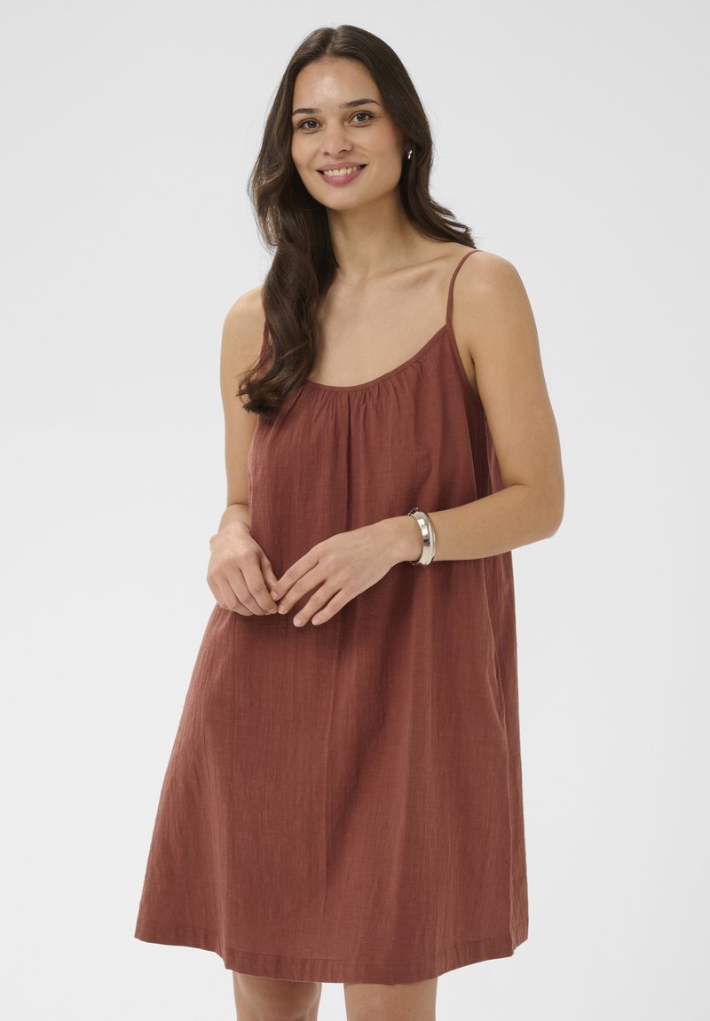 Femme aux longs cheveux bruns portant une robe sans manches couleur rouille et un bracelet argenté, souriante avec les mains doucement jointes.